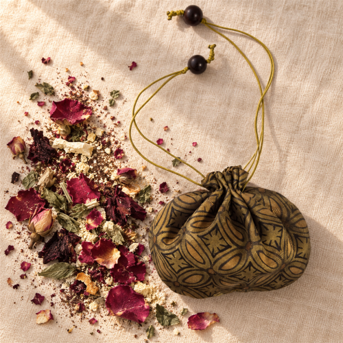 Singular Rose · Carried — sachet and botanicals Herbal sachet in a patterned silk pouch beside dried roses, roselle, mint and crushed peel on linen