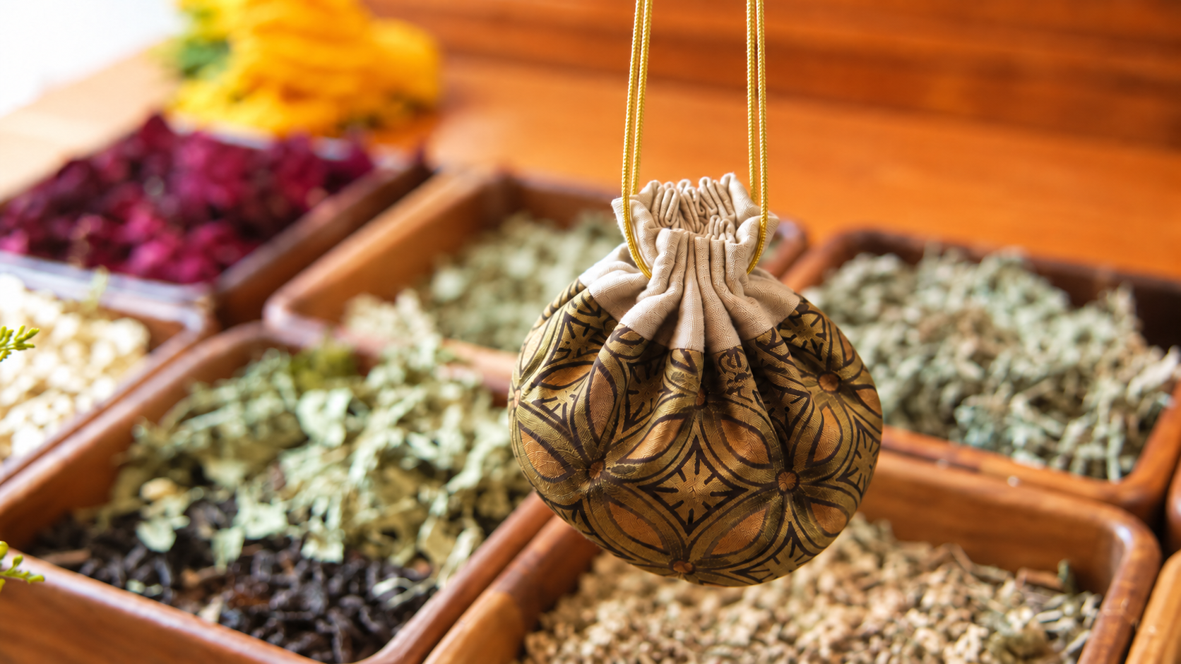 Herbal sachet in a patterned silk pouch hanging on a gold cord above wooden trays of dried herbs
