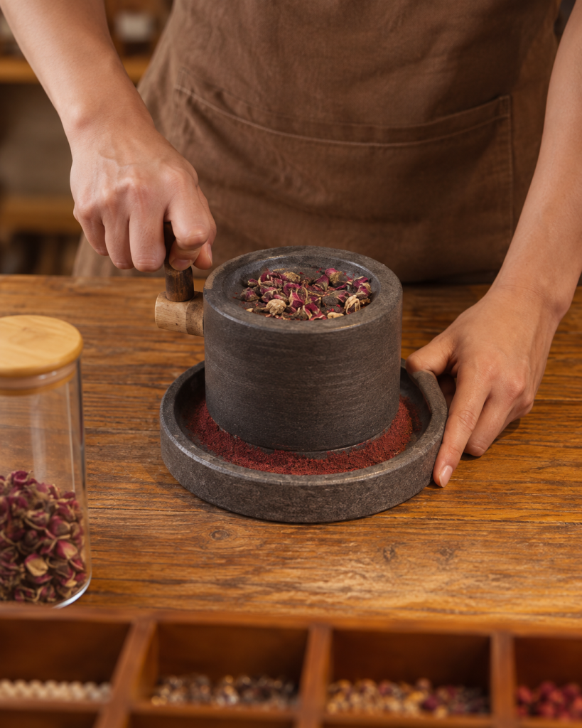 Hands grinding dried rose buds in a dark stone mill on a wooden table