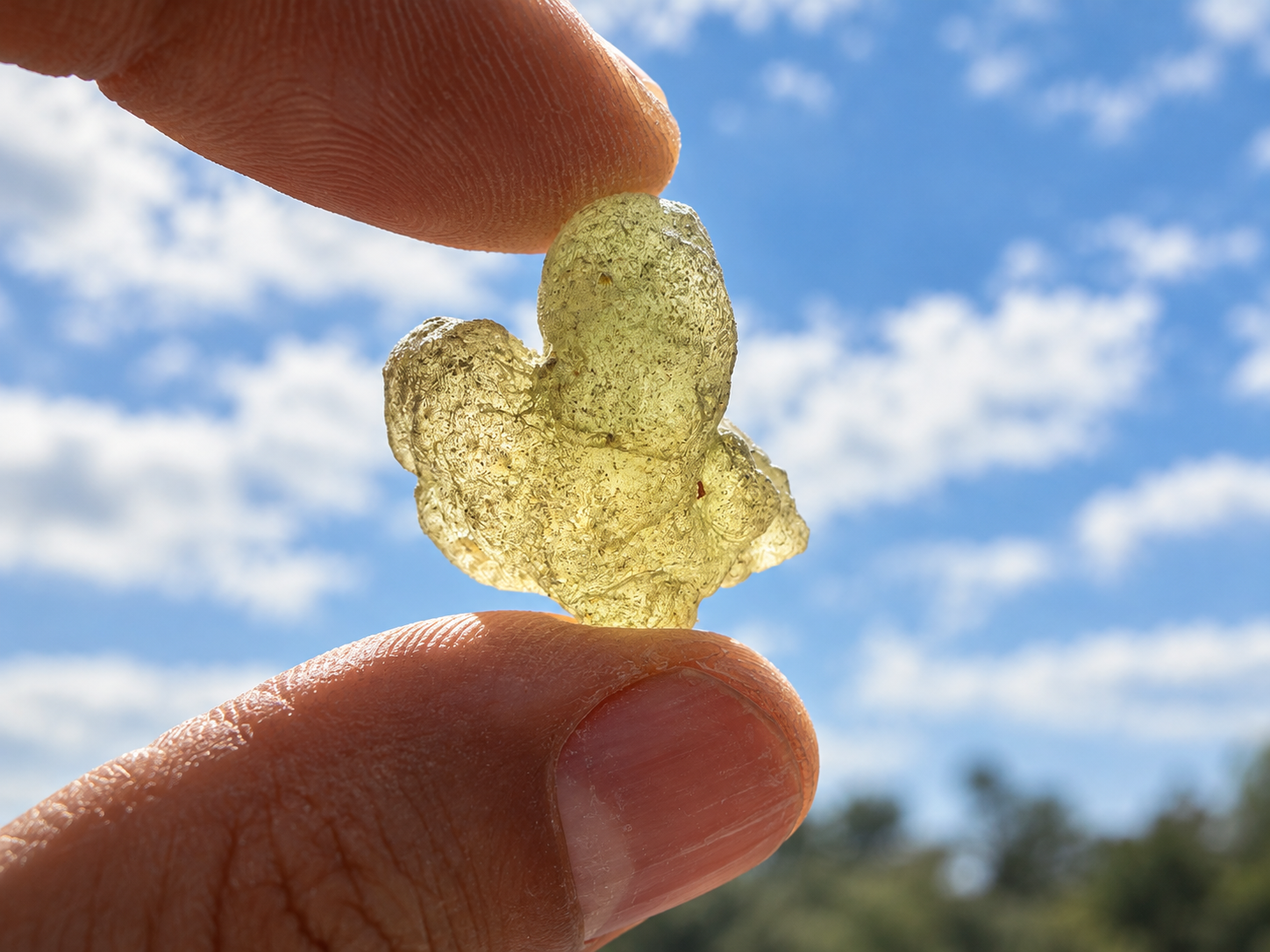 Fingers hold up a pale green piece of frankincense resin against a blue sky with clouds